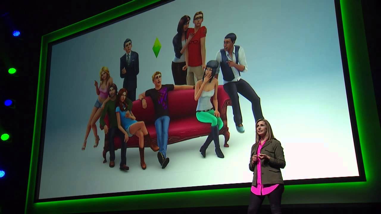 A woman stands on stage in front of a large screen displaying characters from The Sims 4, with several animated characters gathered around a